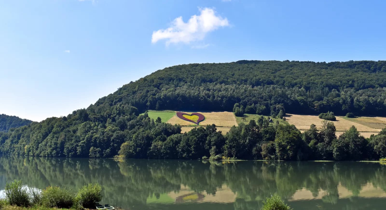 A large yellow heart is carved into a hillside covered in green trees, with a body of water in the foreground and a bright blue sky above.
