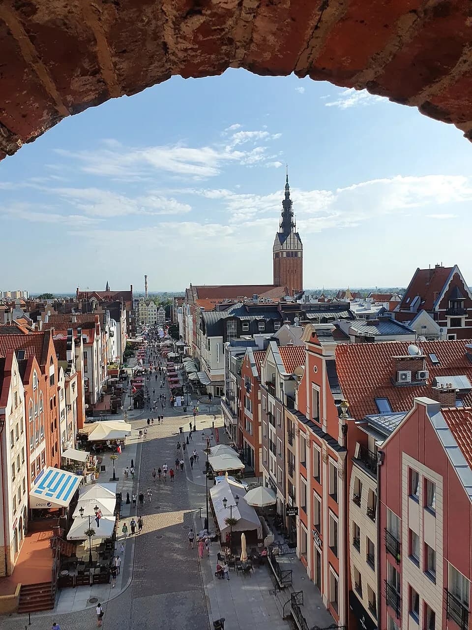 A European town, seen through a brick archway, features a long street lined with buildings leading to a prominent church tower under a blue sky with scattered clouds.